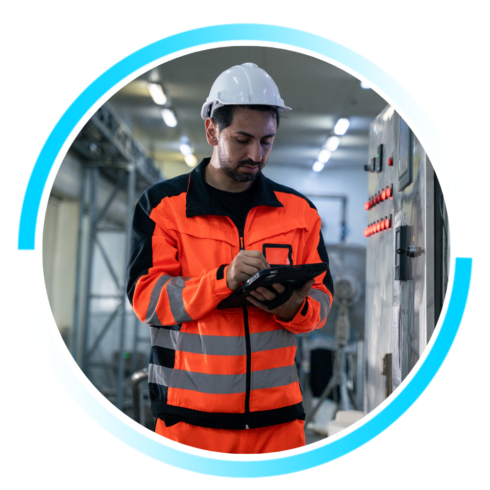 Industrial worker in high-visibility safety gear using a tablet to monitor equipment inside a manufacturing facility.